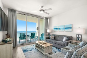 A living room with a view of the ocean, a ceiling fan, and a sliding glass door leading to a balcony.