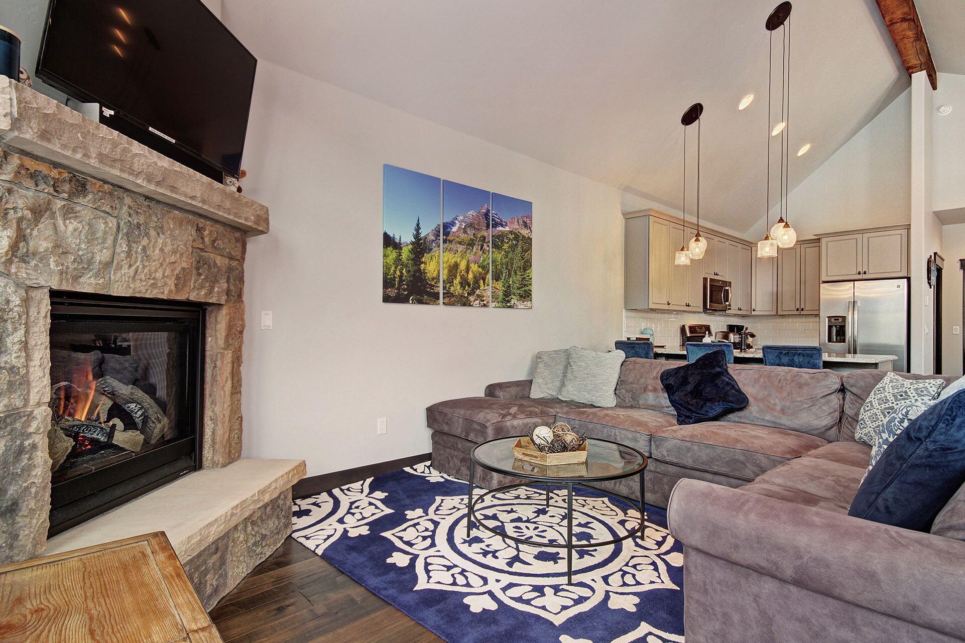 A modern living room with a stone fireplace, a large sectional sofa, a glass coffee table, and a blue patterned rug.