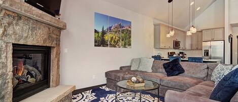 A modern living room with a stone fireplace, a large sectional sofa, a glass coffee table, and a blue patterned rug.