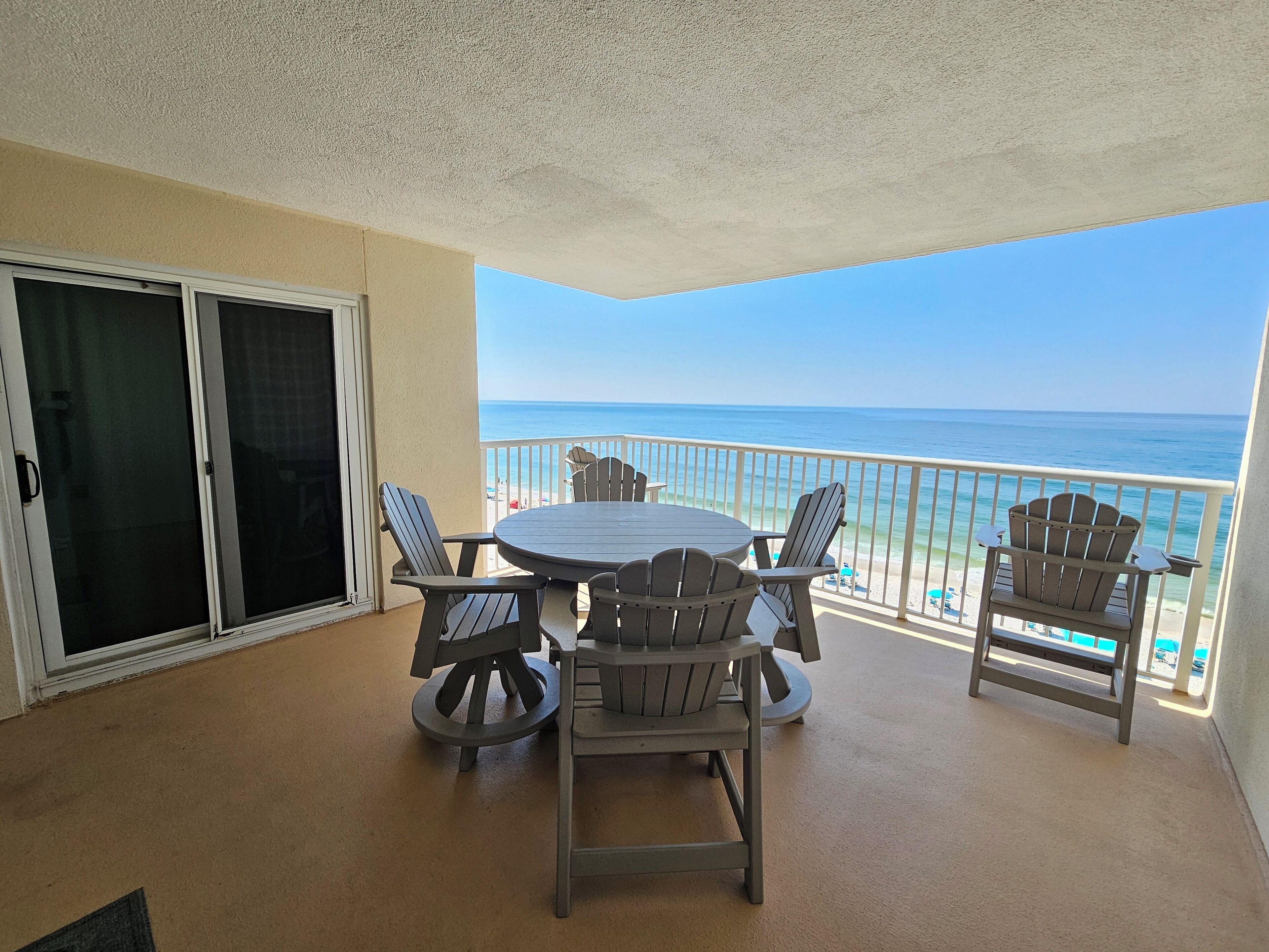 A balcony with a table and chairs, a sliding glass door, and a view of the beach and ocean.