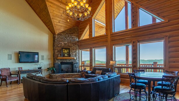 A spacious living room with a stone fireplace, leather seating, a chandelier, and large windows offering a view of the landscape. - The Lodge - Theater Room, Putting Green, Pool, Great view 2 miles to Boone! (Boone)