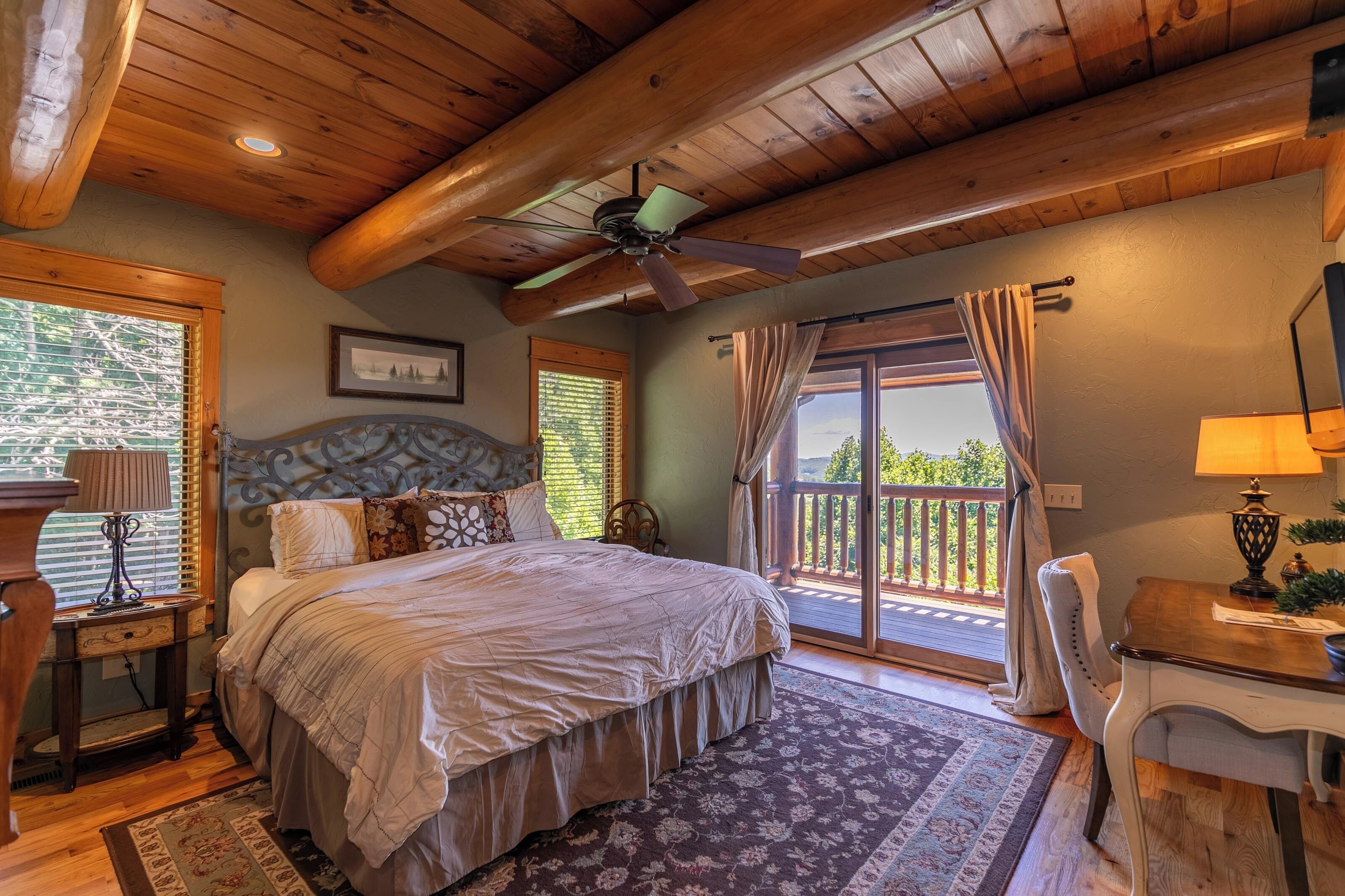 A bedroom with a large bed, a ceiling fan, a wooden ceiling, a window with blinds, a nightstand, a lamp, and a view of the outdoors.