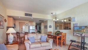 A living room with a sofa, coffee table, and dining area with chairs and a table. There is a kitchen area with cabinets and appliances in the background.