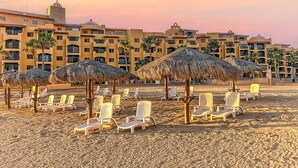 A beach with lounge chairs and thatched umbrellas, a hotel building in the background, and a sunset sky.