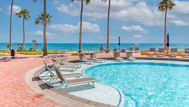 A poolside area with lounge chairs and palm trees, overlooking a clear blue ocean.