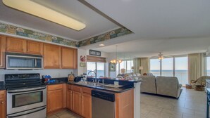 A kitchen with wooden cabinets, a microwave, stove, and dishwasher. There is a dining area with a view of the ocean.