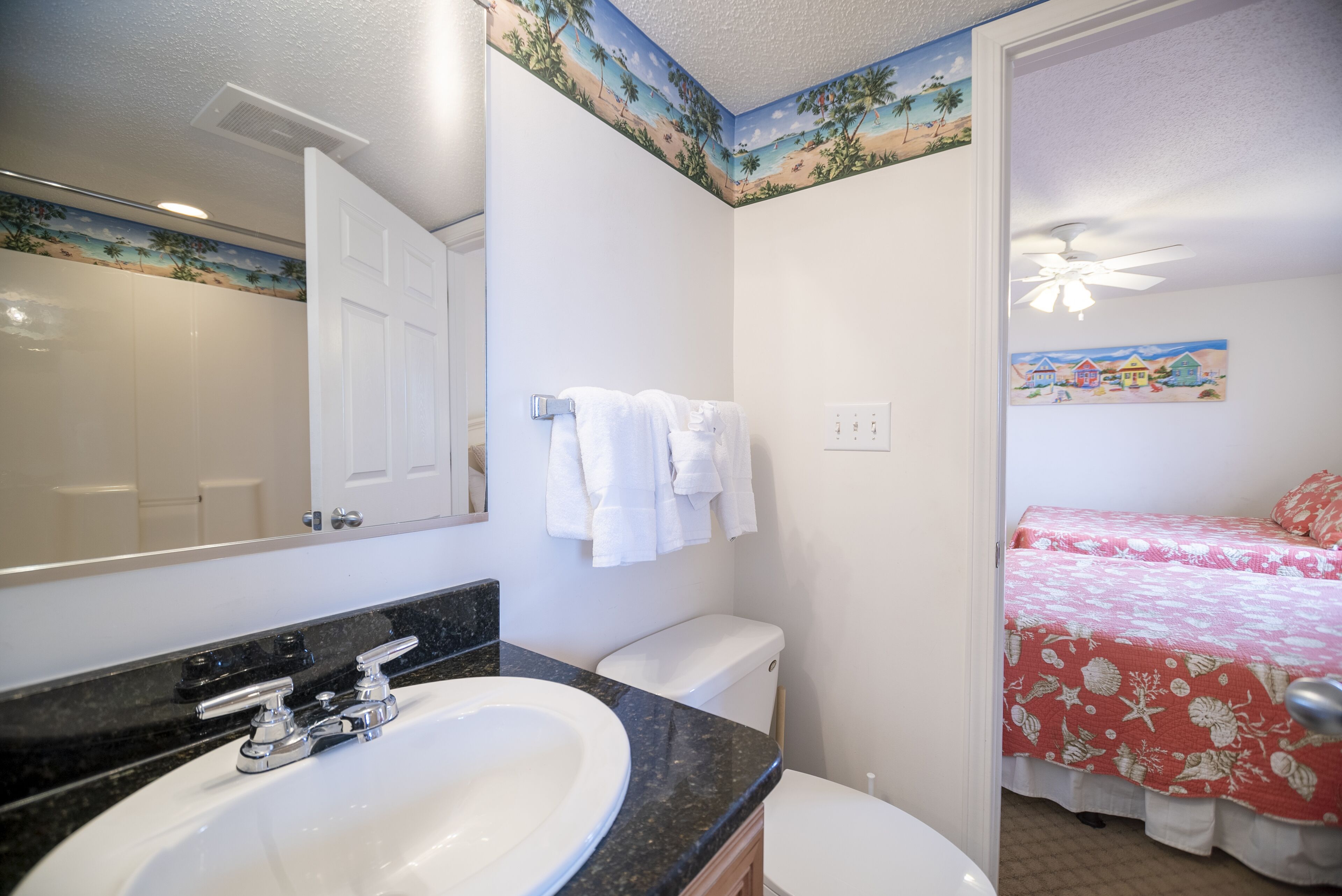 Un baño con lavamanos, inodoro y zona de ducha. Se ve una cama con colcha estampada de flores rojas y blancas a través de una puerta abierta.