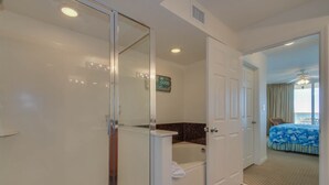 A hotel room with a glass shower enclosure, a bathtub, a bed with a blue floral bedspread, and a view of the ocean through the window.