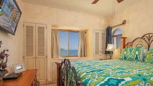 A bedroom with a bed, a desk, a chair, a window with a view of the sea, and a ceiling fan.