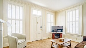 Ein Wohnzimmer mit einem weißen Sessel, einem Holztisch, einem Fernseher auf einem Schrank und einem braunen Sofa.