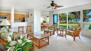 A spacious living area with a dining table, comfortable seating, a ceiling fan, and large windows offering a view of the outdoors. - Kauai Beach Villas F-7  A/C & Oceanfront Lanai (Lihue)