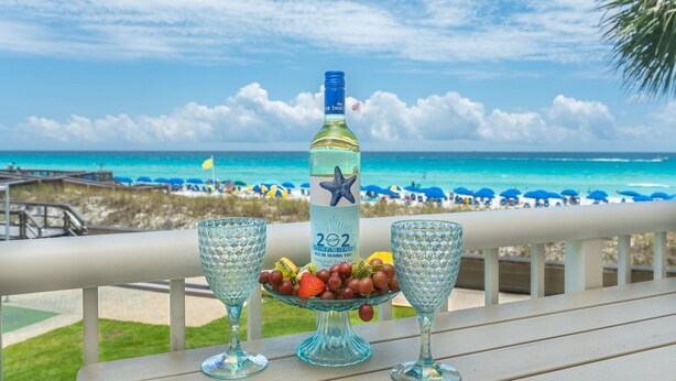 A table with a bottle and two glasses on a balcony overlooking a beach with umbrellas and people. - 202- Newly renovated upscale Gulf Front Condo, Includes FREE Beach Service (Destin)