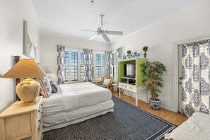 Ein Schlafzimmer mit Bett, Schreibtisch, Stuhl, Fernseher, einer Pflanze und einem Deckenventilator.