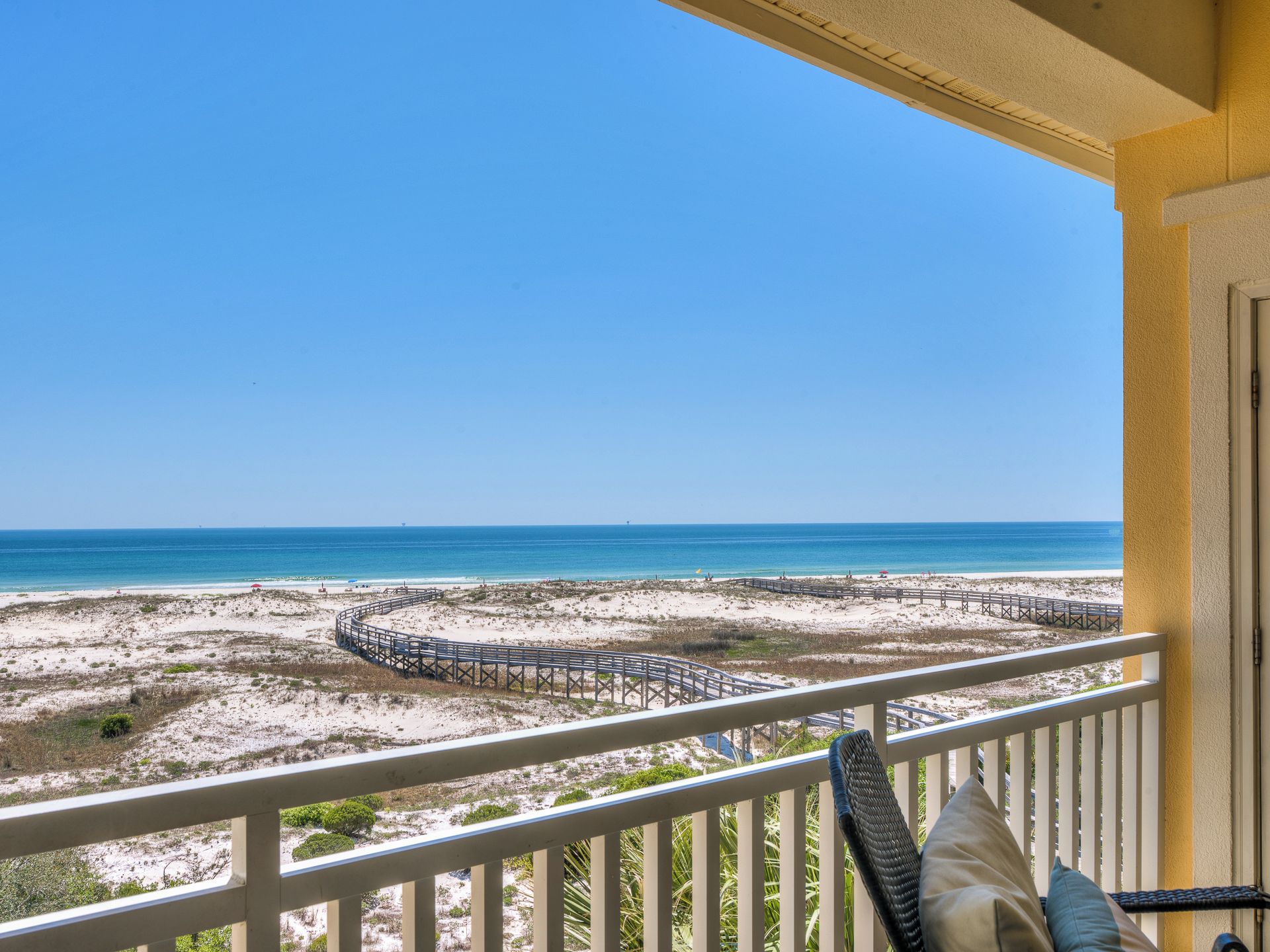 A balcony with a view of a beach, a wooden boardwalk, and a clear blue sky.