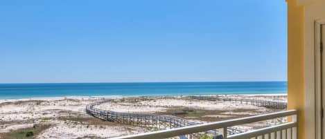 A balcony with a view of a beach, a wooden boardwalk, and a clear blue sky.