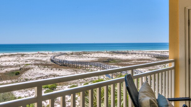 Ein Balkon mit Blick auf einen Strand, eine Holzpromenade und einen wolkenlosen, blauen Himmel.