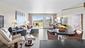 A modern living room with a sofa, chairs, a coffee table, and a dining area with a table and chairs. There is a television and a lamp on a side table. - 120- Walkout Balcony to Pool or Beach, Entirely Remodeled, FREE beach service (Destin)