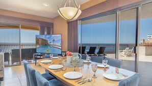 Zona pranzo con vista sull'oceano, una televisione che mostra una località turistica e un tavolo apparecchiato per quattro persone.