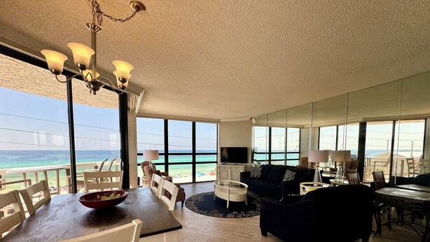 A spacious living room with a large window overlooking the ocean, a dining area with a wooden table and chairs, and a cozy seating area with a dark-colored sofa and armchairs. - 2 Free Beach SRVCS- 4 chairs/2 umbrellas! Free Beach Bonfires M/F nts (Miramar Beach)