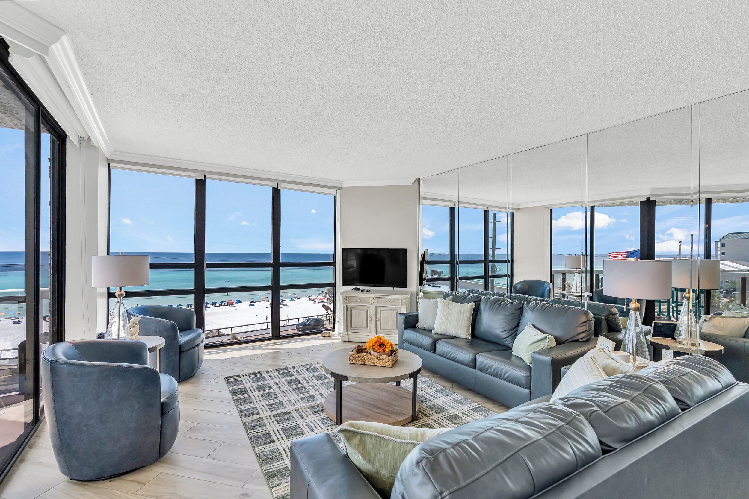 A modern living room with a large window overlooking a beach, a flat-screen TV, and a comfortable seating area.