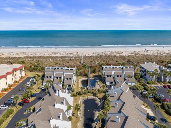 A coastal hotel with multiple buildings, a parking lot, and a beach view.