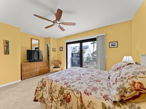 Une chambre à coucher avec un lit, un ventilateur de plafond, un téléviseur posé sur une commode en bois, une chaise et un balcon offrant une vue sur les bâtiments et les arbres.