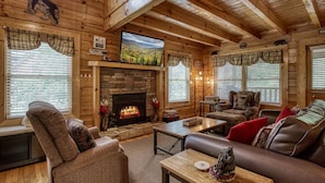 A cozy cabin interior with a stone fireplace, wooden walls, and a large window overlooking a scenic landscape. - ERN834 - LAKESIDE GETAWAY - GREAT LOCATION!  CLOSE TO ALL THE ACTION! (Pigeon Forge)