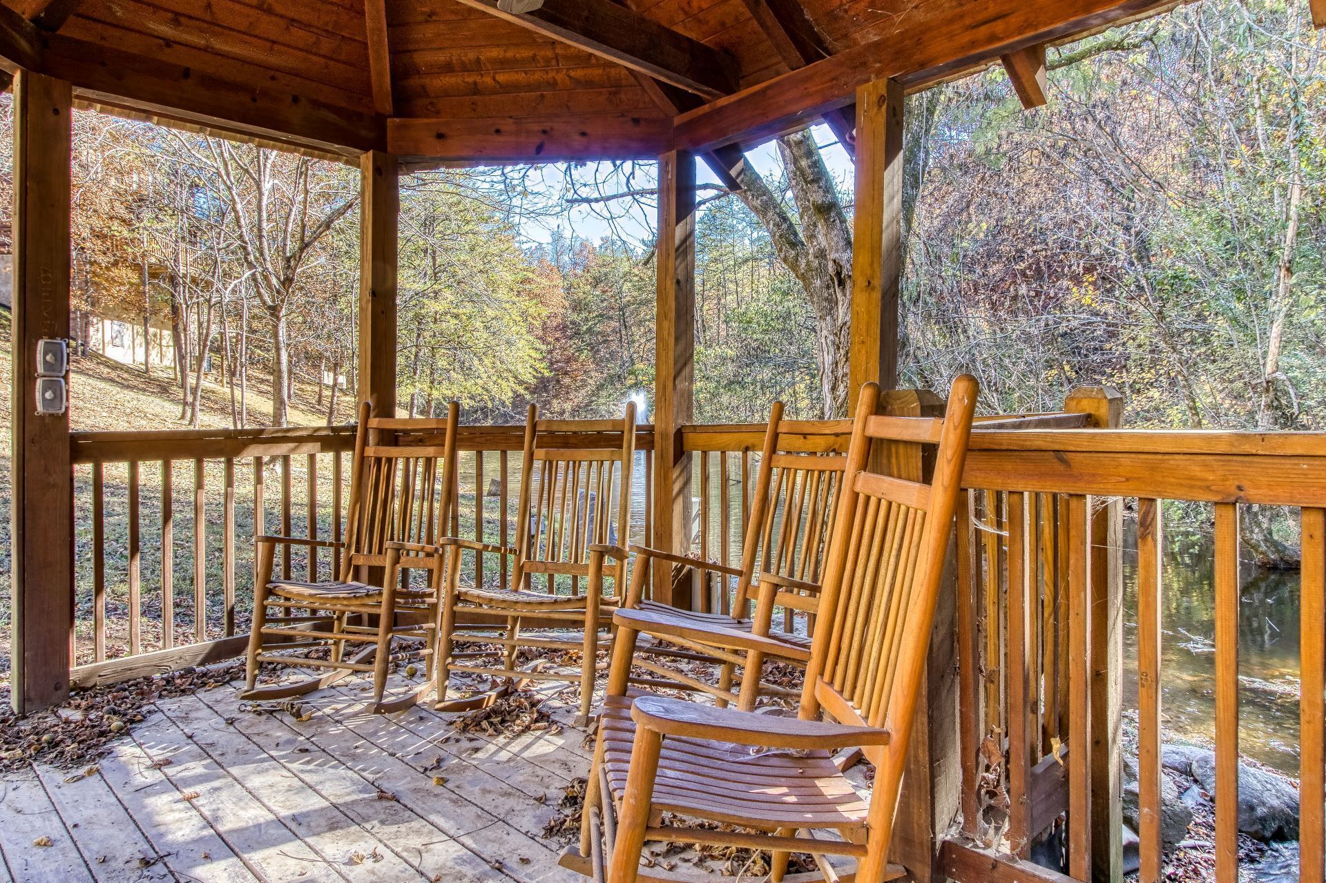 A covered wooden deck with several wooden chairs and a railing overlooking a forested area.