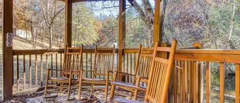 A covered wooden deck with several wooden chairs and a railing overlooking a forested area.