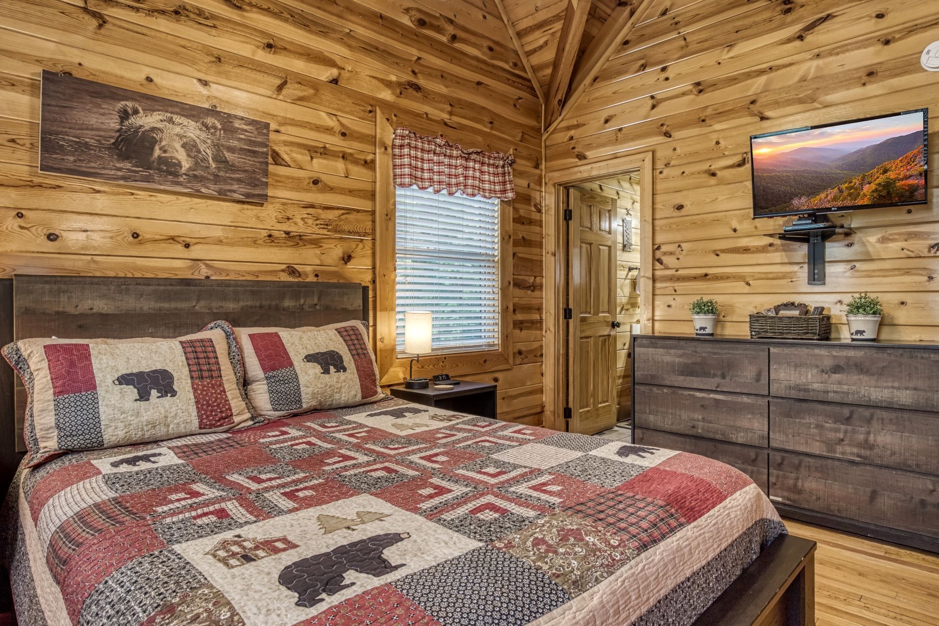 A log cabin bedroom with a large bed, a wooden headboard, a wooden dresser, a television, and a window with blinds.
