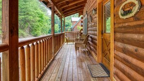 A wooden cabin with a porch featuring wicker chairs, a welcome sign, and a view of the forest. - ERN834 - LAKESIDE GETAWAY - GREAT LOCATION!  CLOSE TO ALL THE ACTION! (Pigeon Forge)