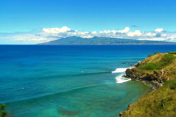 A coastal landscape with clear blue waters, a rocky shoreline, and a mountain in the distance.