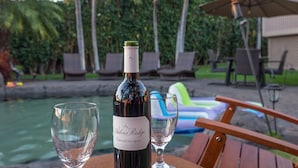 Uma garrafa de vinho e duas taças sobre uma mesa, perto de uma piscina com espreguiçadeiras e um guarda-sol ao fundo.