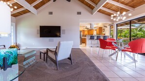 Uma sala de estar ampla com teto alto, uma televisão na parede e uma cozinha visível através de uma porta aberta.