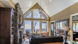 A spacious living room with a large window, a stone fireplace, and a cozy seating area. - View House  - Stunning Views of Mountains and Yadkin River, huge deck, wood burning fireplace (Boone)