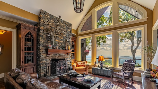 A spacious living room with a stone fireplace, leather sofa, and large windows offering a view of the outdoors. - View House  - Stunning Views of Mountains and Yadkin River, huge deck, wood burning fireplace (Boone)