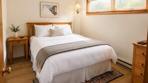 Quarto com cama, mesas de cabeceira, um criado-mudo, luminária, janela com persianas e um quadro na parede.