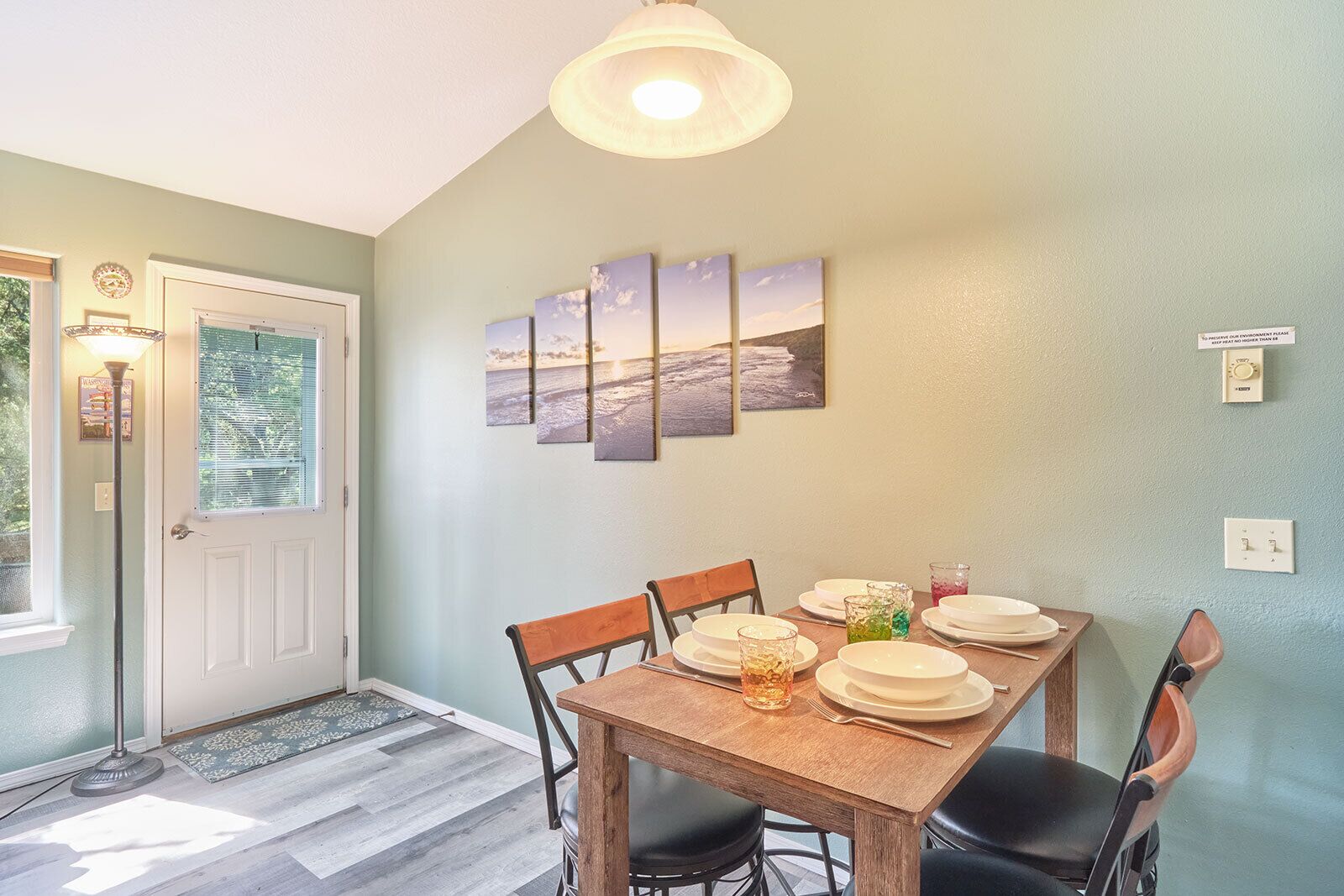 Un coin repas avec une table en bois dressée pour quatre personnes, une fenêtre avec des stores et une œuvre d’art accrochée au mur représentant un coucher de soleil sur l’eau.