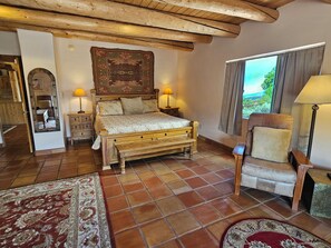 A bedroom with a wooden bed, two bedside lamps, a large window with curtains, and a view of greenery outside.