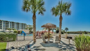 A resort pool area with palm trees, lounge chairs, and a thatched umbrella. - The Islander 110 - Winter Months Available - Heated Pool (Destin)