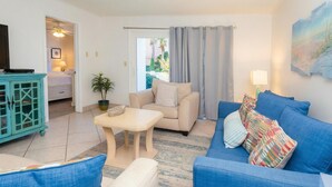A living room with a beige sofa, a blue sofa, a small table, a teal cabinet, a TV, and a window with curtains. - The Islander 110 - Winter Months Available - Heated Pool (Destin)