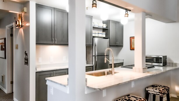 A modern kitchen with grey cabinets, a stainless steel refrigerator, a sink, and a microwave. There are two stools with a leopard print design. - JHRL - Balsam Lodge A8 - Great Location in the Aspens (Wilson)