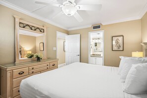 Une chambre avec un grand lit, une commode avec des tiroirs, un miroir, un ventilateur de plafond et une porte donnant sur une autre pièce.