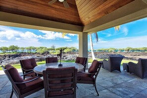 Un espace extérieur aménagé, abrité et doté d’une table ronde et de chaises, offrant une vue panoramique sur un paysage verdoyant, avec des arbres et la mer à l’horizon.