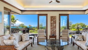 A spacious living area with a glass coffee table, two beige armchairs, and a sofa with patterned cushions. Large windows offer a view of a tropical landscape.