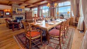 A spacious dining area with a wooden table set for six, surrounded by chairs with patterned cushions. A stone fireplace and a television are visible in the background. - JHRL - Amazing 5-bedroom Granite Ridge Lodge #16, Hot Tub and Sweeping Views (Teton Village)