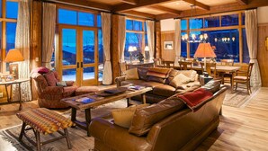 A cozy living room with a leather sofa, a red armchair, and a dining table with chairs. Large windows offer a view of the outdoors. - JHRL - Amazing 5-bedroom Granite Ridge Lodge #16, Hot Tub and Sweeping Views (Teton Village)