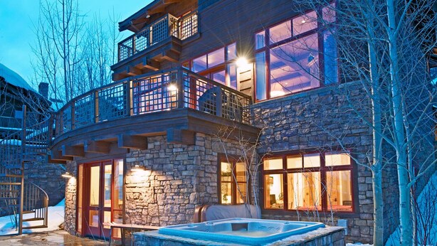 A multi-story stone building with a stone staircase, large windows, and a hot tub in the snow. - JHRL - Amazing 5-bedroom Granite Ridge Lodge #16, Hot Tub and Sweeping Views (Teton Village)
