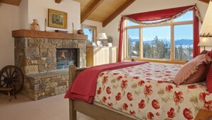 A bedroom with a stone fireplace, a bed with floral bedding, a wooden headboard, and a view of mountains through a large window. - JHRL - Amazing 5-bedroom Granite Ridge Lodge #16, Hot Tub and Sweeping Views (Teton Village)