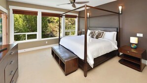 A four-poster bed with a wooden headboard, a bench at the foot of the bed, a nightstand with a lamp, and large windows with a view of greenery. - Nature's Gate Executive Estate (Victoria)